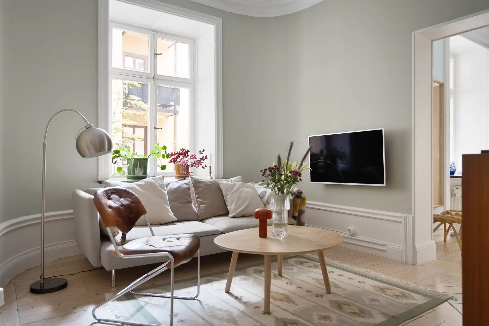 Šviesus atnaujintas buto kambarys su sofa, aukštais langais ir šviesiomis medinėmis grindimis